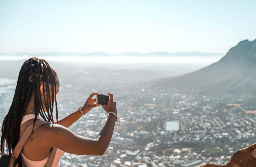 A woman with braided hair takes a photo with her smartphone from a high vantage point overlooking a city, with mountains in the background.