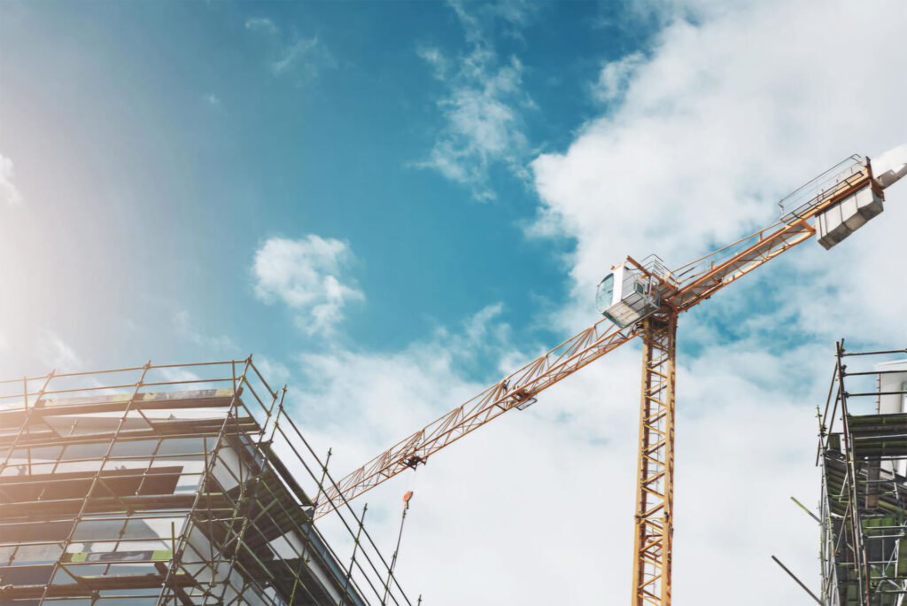 A construction crane towering above a building under construction, set against a bright blue sky with clouds.